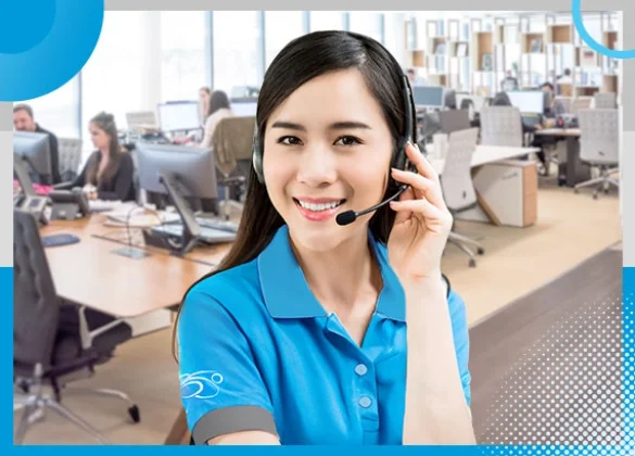 Smiling customer support representative wearing a headset and a blue uniform, standing in a modern office environment with desks and computers in the background.