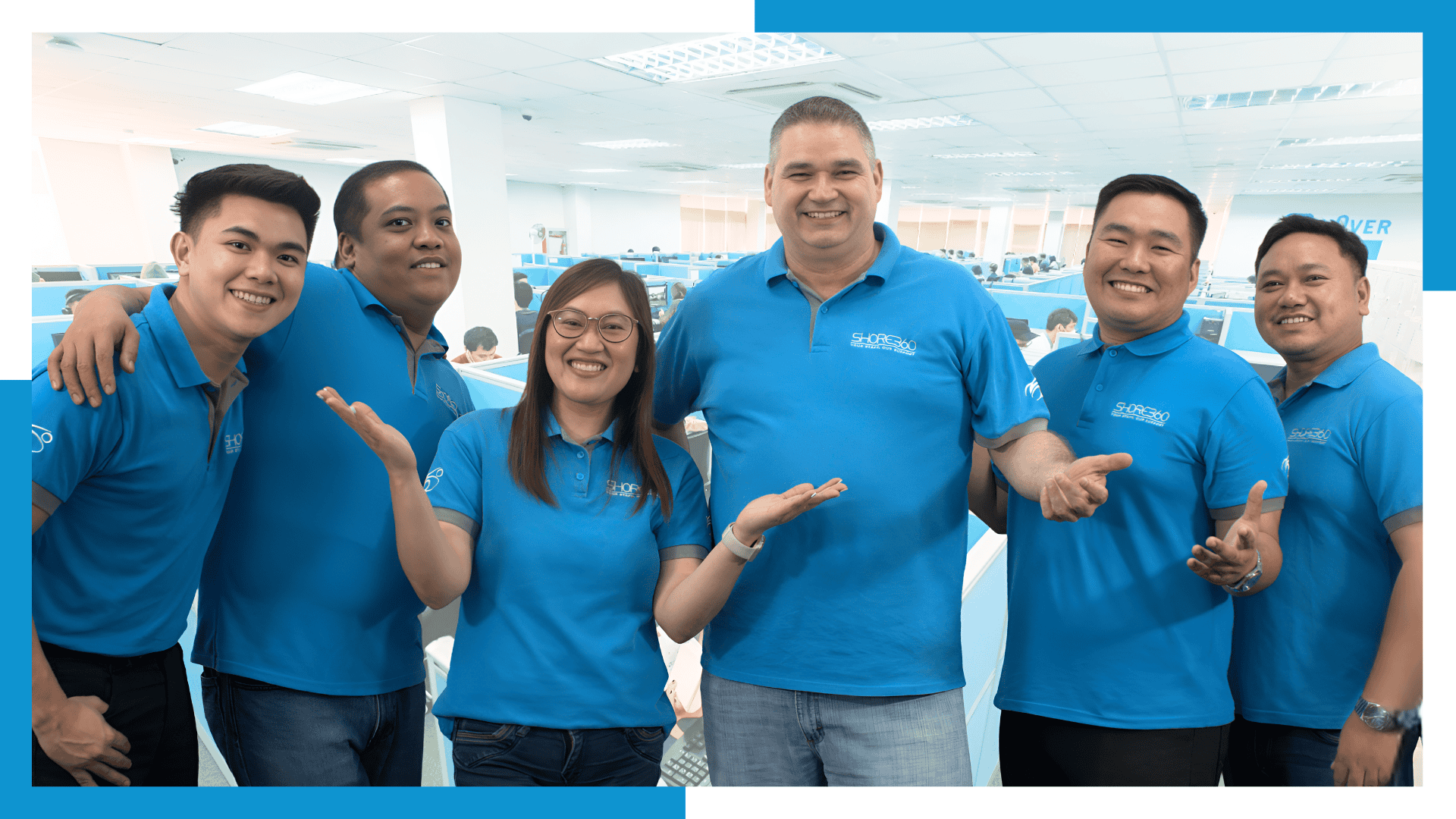 Diverse and cheerful offshore team members in blue Shore360 shirts, including Filipino and Western professionals, smiling and collaborating in a modern office setting in the Philippines.