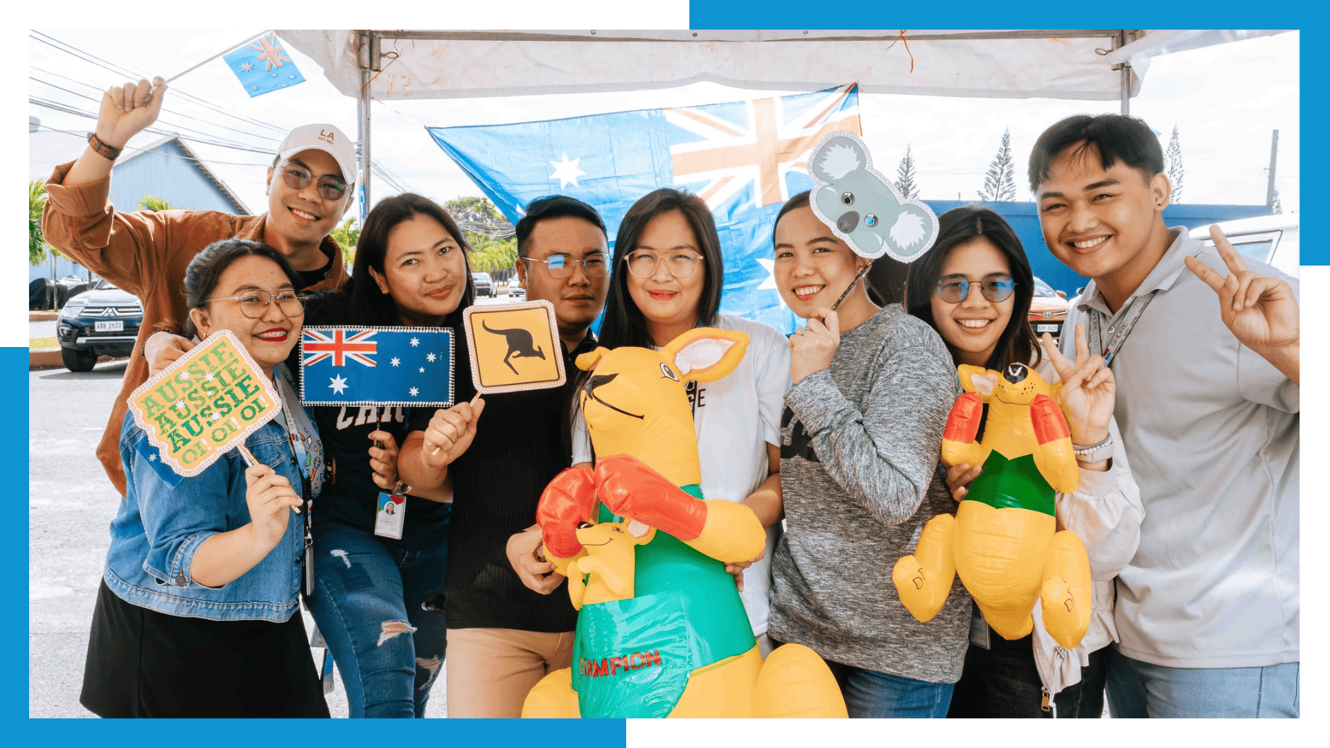 A joyful group of young Filipino professionals, holding Australian-themed props like an inflatable kangaroo, koala headbands, and signs, celebrating a company event. An Australian flag is visible in the background.