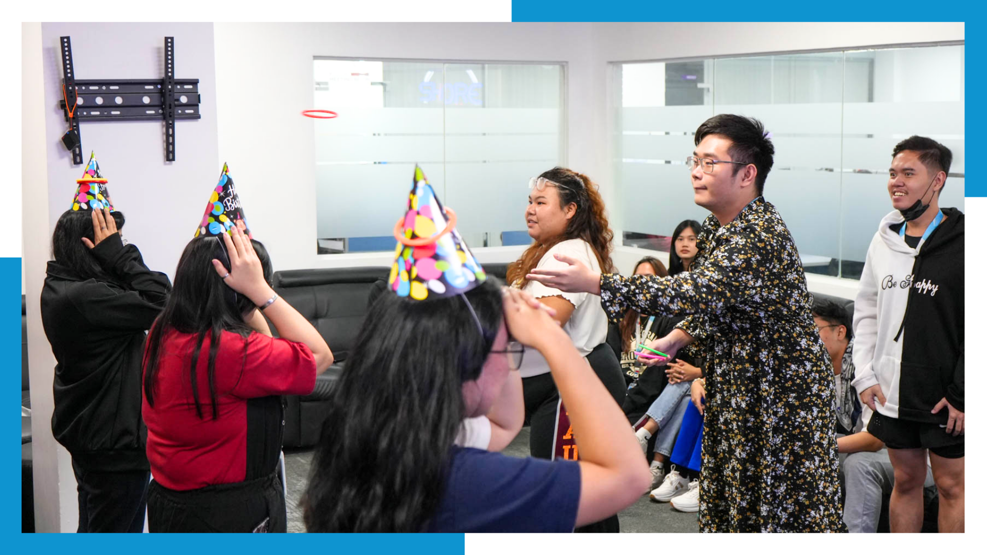 A cheerful group of young Filipino professionals, wearing colorful birthday party hats, are celebrating in a modern office space. One person in the foreground is putting on a hat, while others in the background are smiling and talking, creating a lively and festive atmosphere.