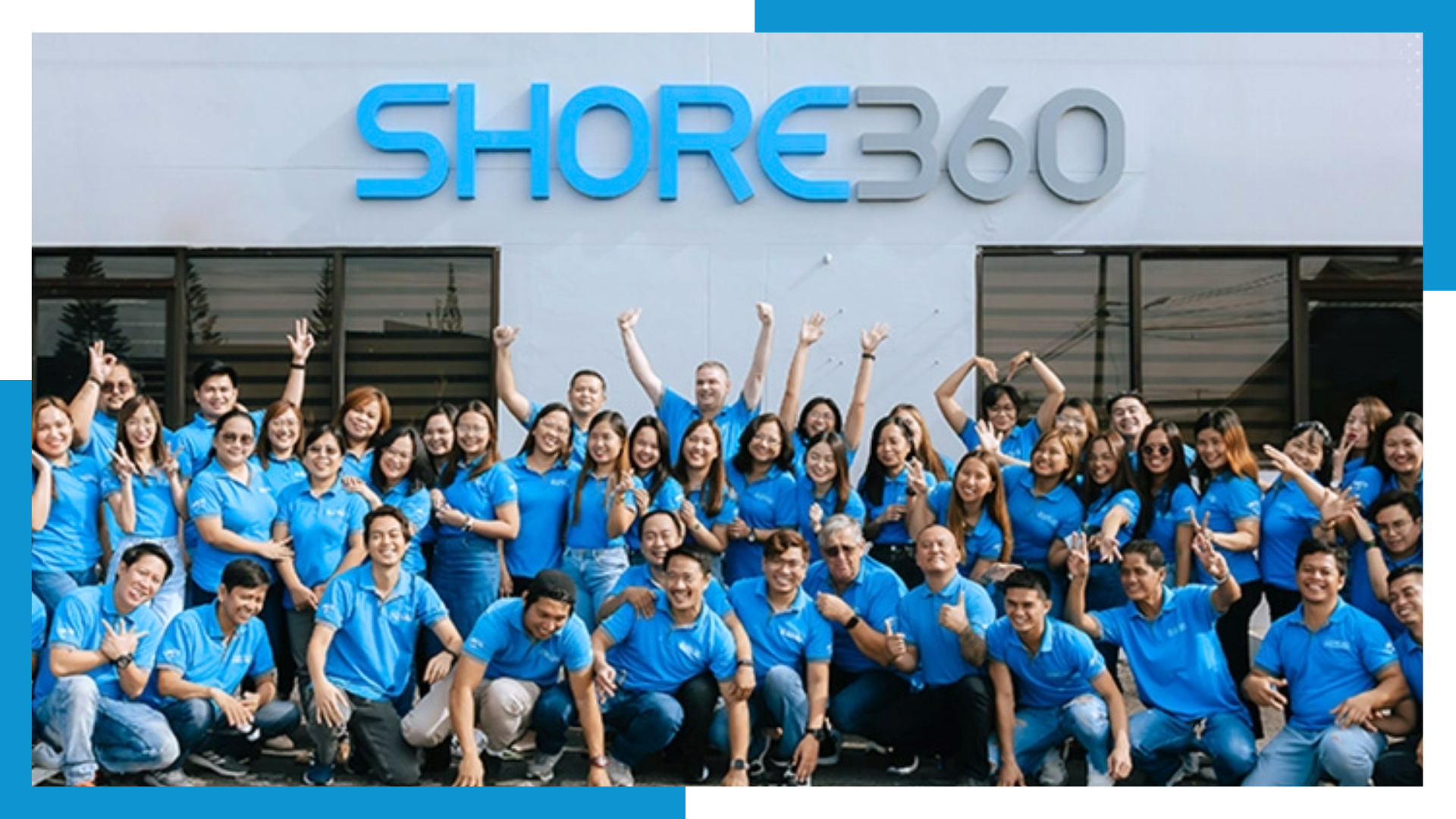 A large and diverse group of Filipino professionals wearing blue Shore360 shirts. They are smiling, waving, and posing enthusiastically in front of the Shore360 building in the Philippines, showcasing a vibrant company culture and a strong, united team.